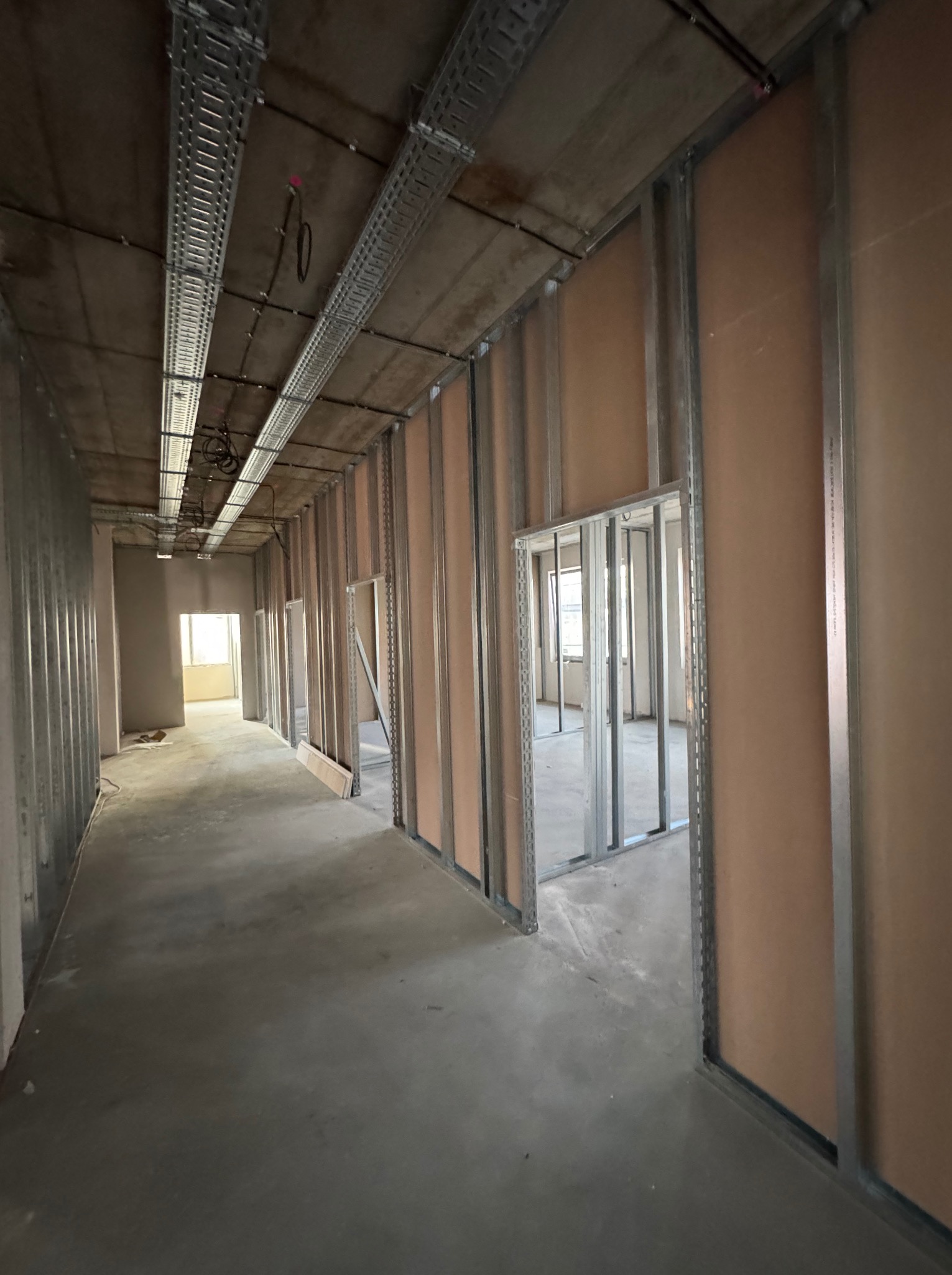 Wnętrze w trakcie prac: widok na korytarz z metalową konstrukcją ścian działowych i częściowo zamontowanymi płytami. Widoczne koryta kablowe pod sufitem.