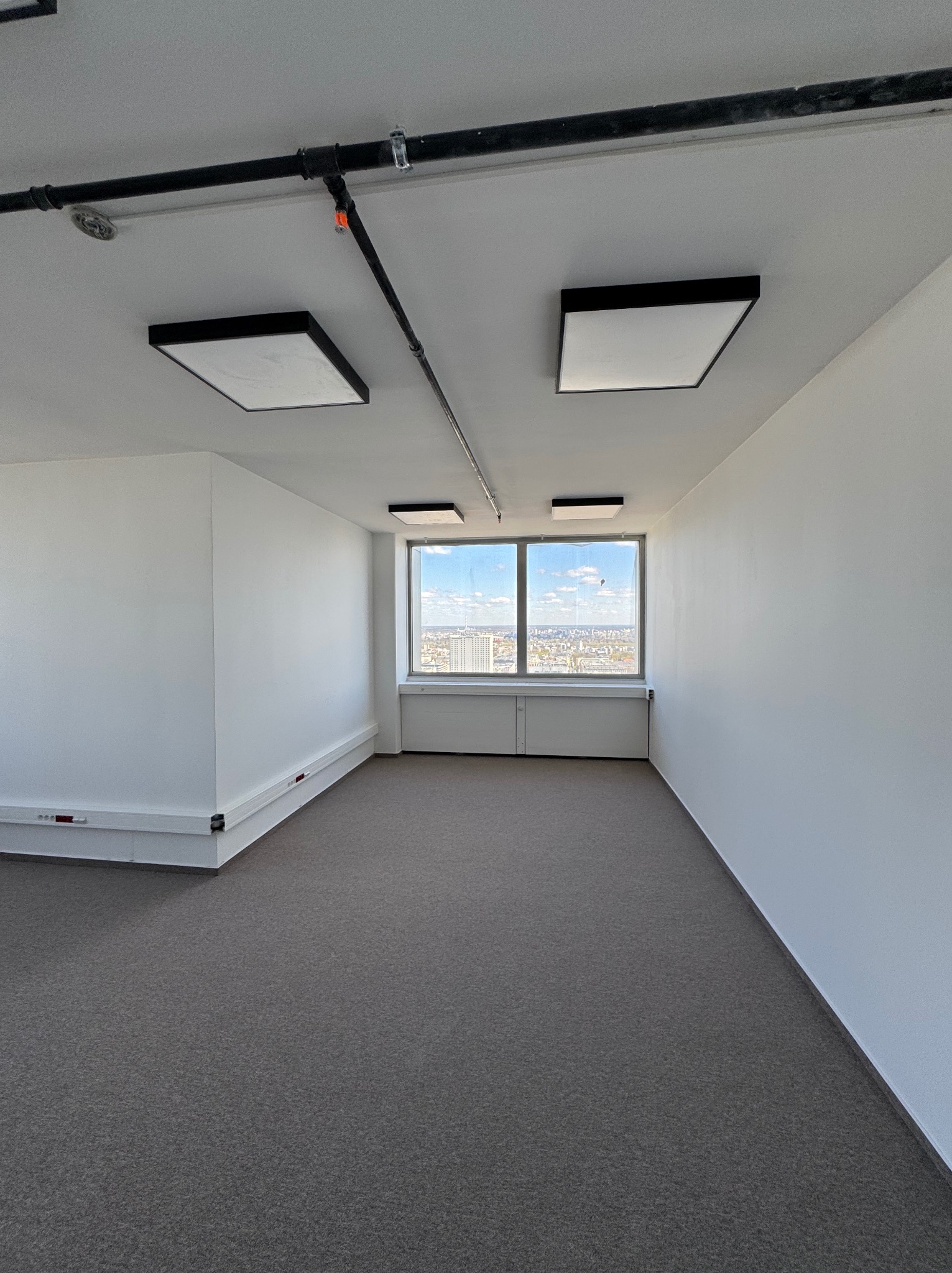 Jasne, odnowione biuro z szarym dywanem, białymi ścianami i oknem z widokiem na miasto. Widoczne czarne rury pod sufitem i nowoczesne, kwadratowe lampy.