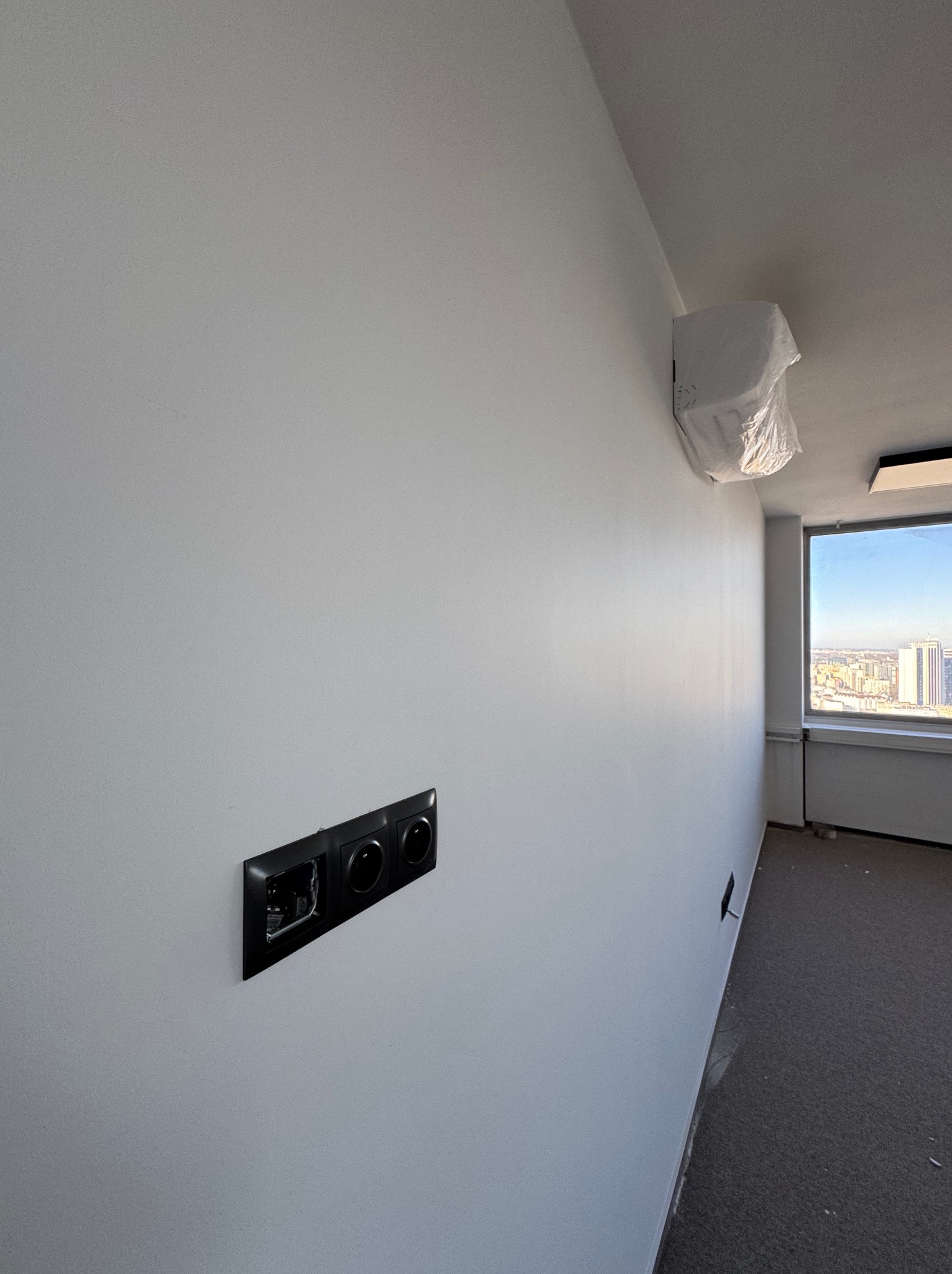 Świeżo pomalowana ściana w biurze z gniazdkami elektrycznymi, klimatyzacja w folii ochronnej i widokiem na miasto z okna. Jasne wnętrze po odnowieniu.