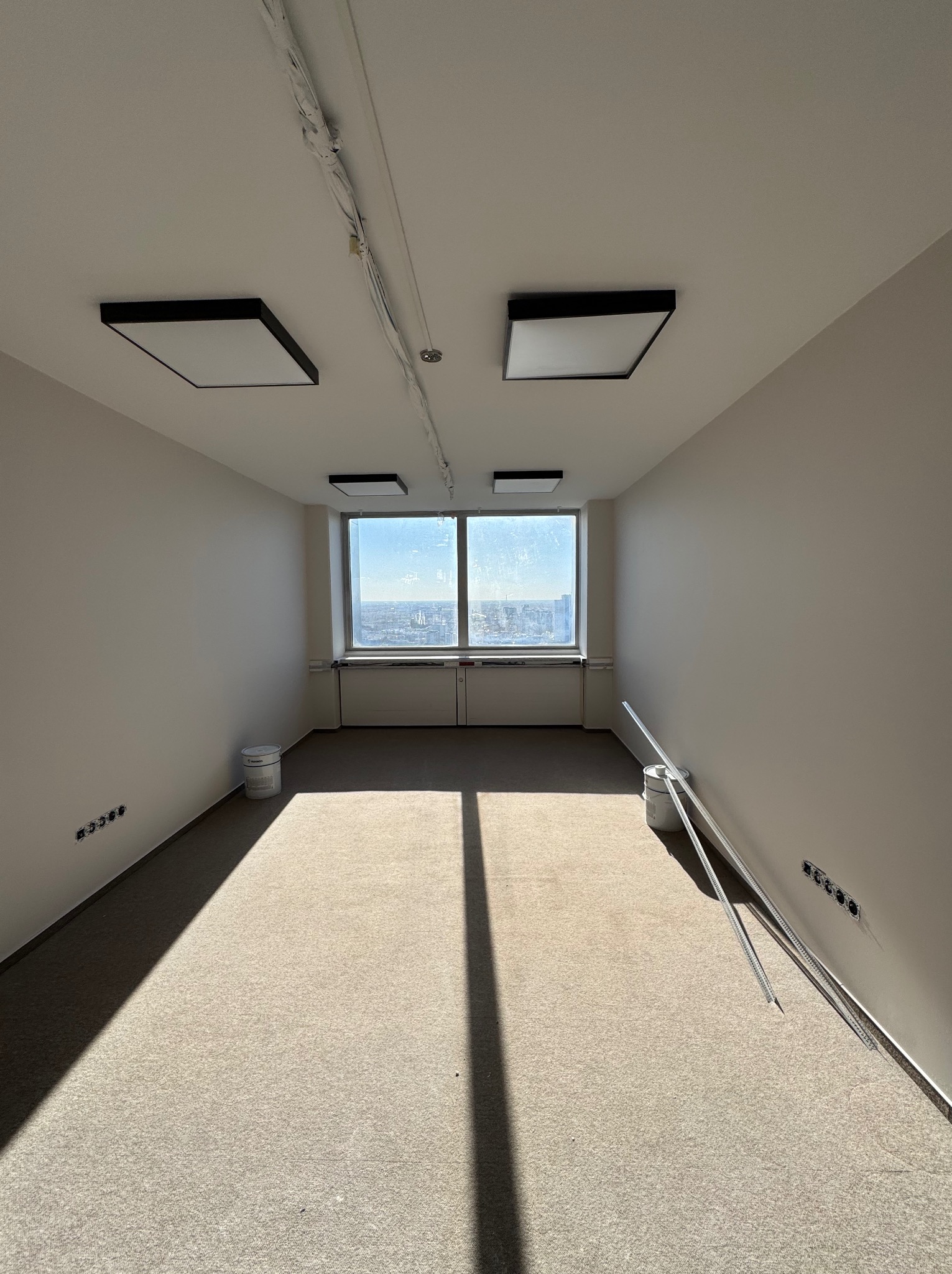 Wnętrze biura po odświeżeniu: jasne ściany, okno z widokiem na miasto, nowoczesne oświetlenie sufitowe. Widoczne materiały budowlane, sugerujące etap wykończeniowy.