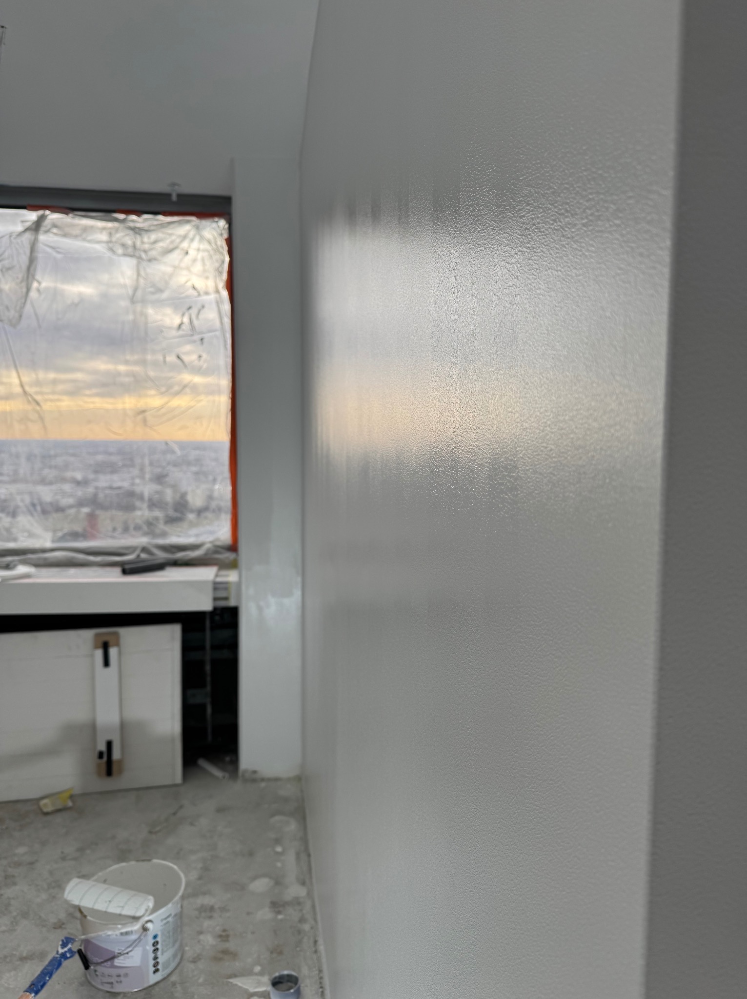 Świeżo pomalowana, gładka ściana w odcieniach szarości w pokoju z widokiem na miasto, z wiaderkiem farby i wałkiem na podłodze. Okno zabezpieczone folią malarską.