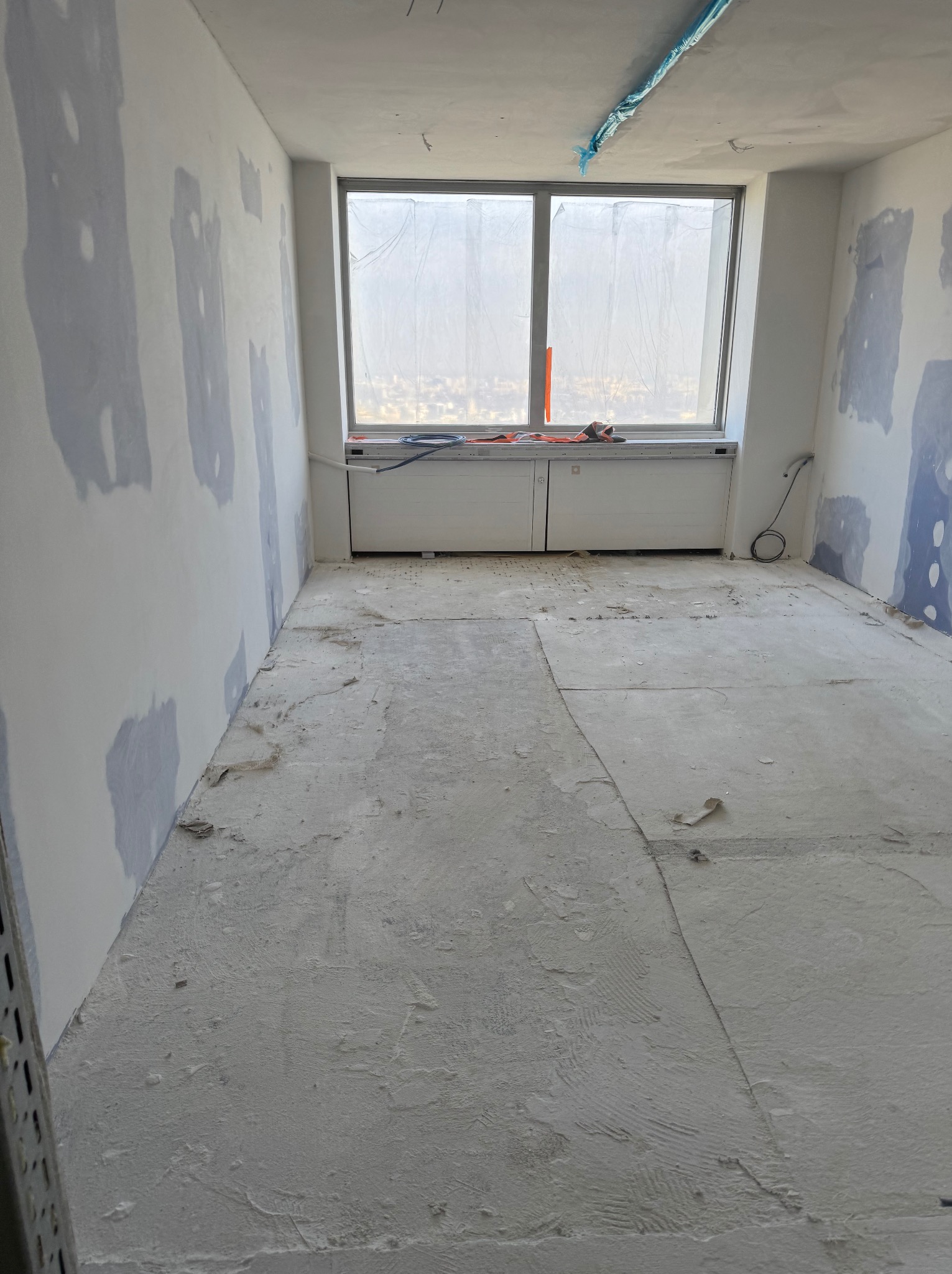 Pokój w trakcie prac wykończeniowych: częściowo wyszpachlowane ściany z widocznymi łatami, surowa podłoga, okno z widokiem na miasto w tle.