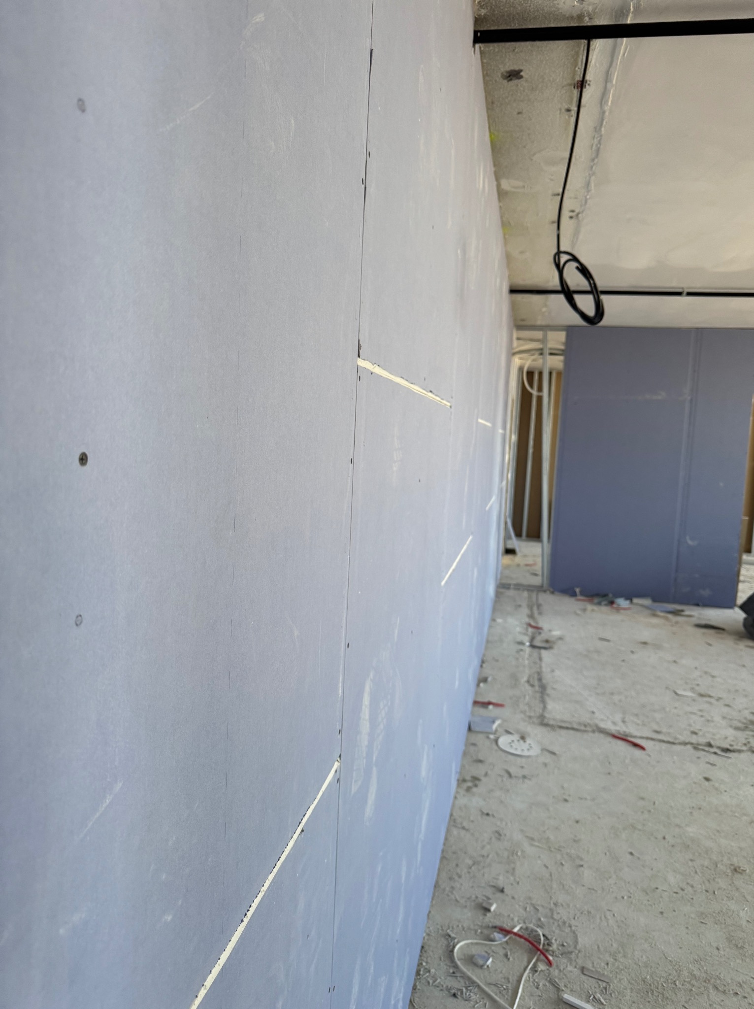 Ściana z płyt kartonowo-gipsowych w trakcie montażu, widoczne łączenia i surowy stan wykończenia wnętrza. W tle instalacje i przewody.