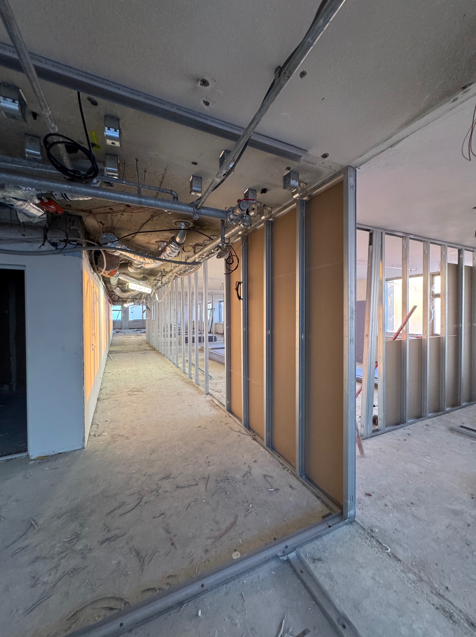 Wnętrze w trakcie budowy, widoczne metalowe profile ścian działowych i częściowo zamontowane płyty. Instalacje pod sufitem, surowa podłoga. Perspektywa długa, wąska.