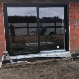 Montaż czarnego okna przesuwnego w surowym murze z czerwonej cegły, widoczny fragment drabiny i folia ochronna na dole, w szybie odbija się sylwetka osoby i budynek w tle.