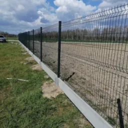 Długi fragment panelowego ogrodzenia z podmurówką betonową na trawiastym terenie, widok z boku w słoneczny dzień.