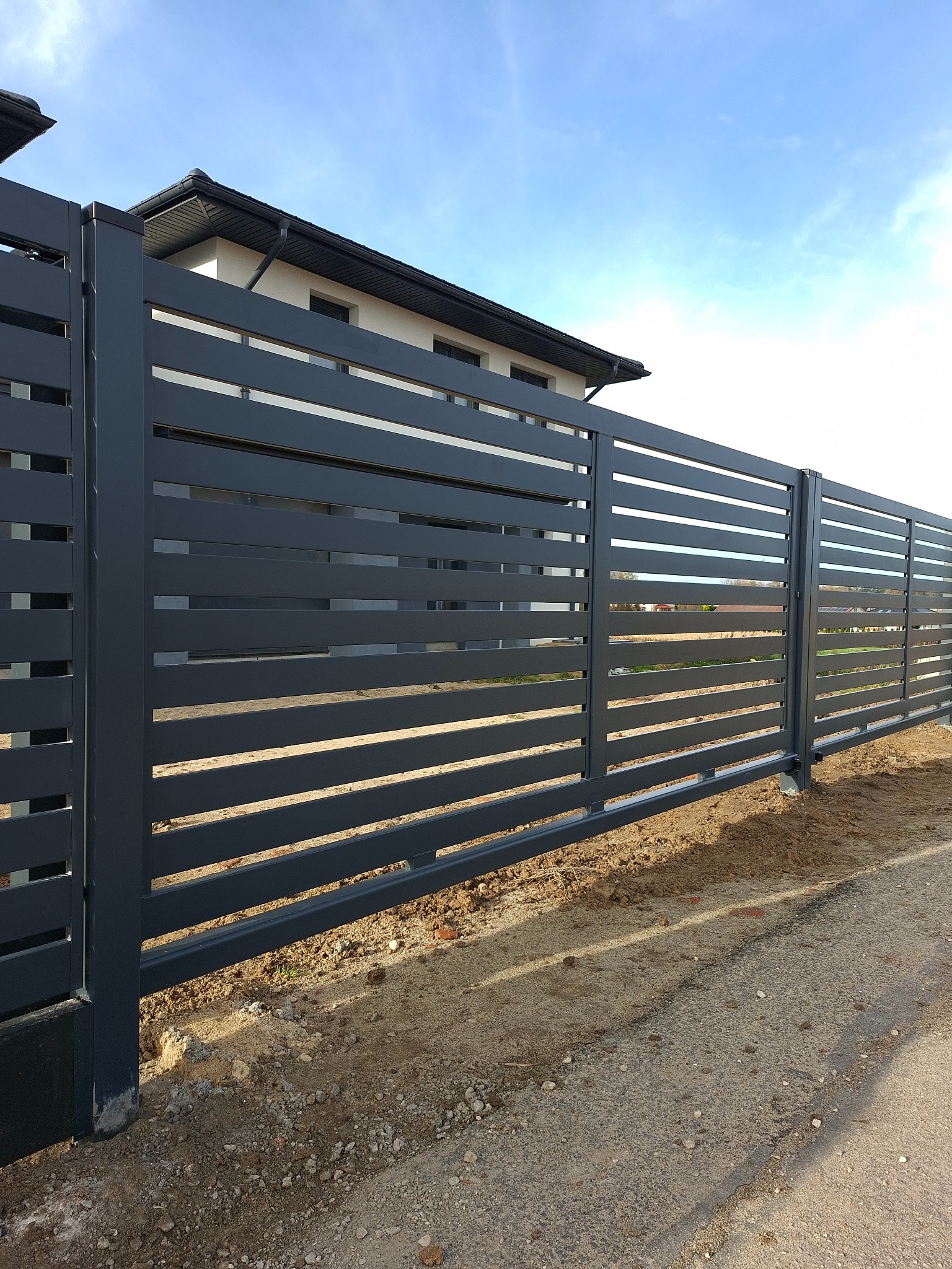 Nowoczesne, poziome ogrodzenie panelowe w kolorze grafitowym, z widocznym fragmentem domu w tle, na tle błękitnego nieba. Minimalistyczny design.