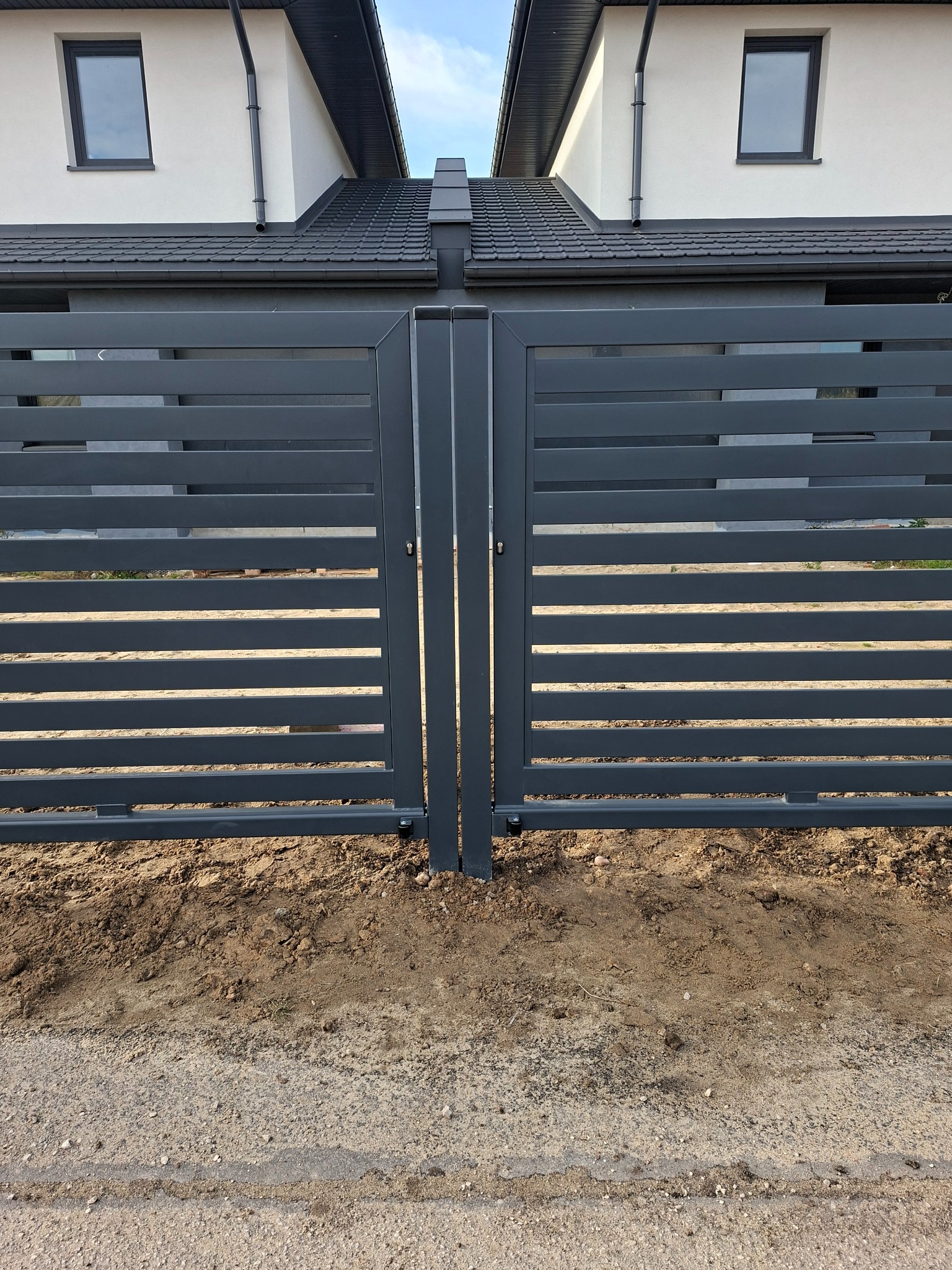 Nowoczesna, pozioma, metalowa brama w kolorze grafitowym, z widocznymi dwoma budynkami w tle. Minimalistyczny design, świeżo zamontowana, z ziemią u podstawy.