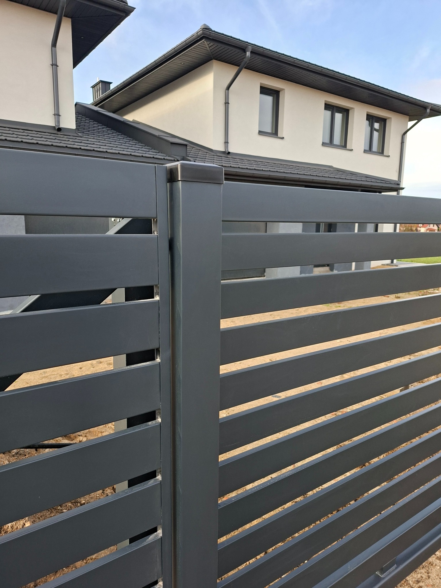 Nowoczesne, poziome ogrodzenie panelowe w kolorze grafitowym, z widocznym fragmentem domu w tle. Minimalistyczny design, równoległe panele, precyzyjny montaż.