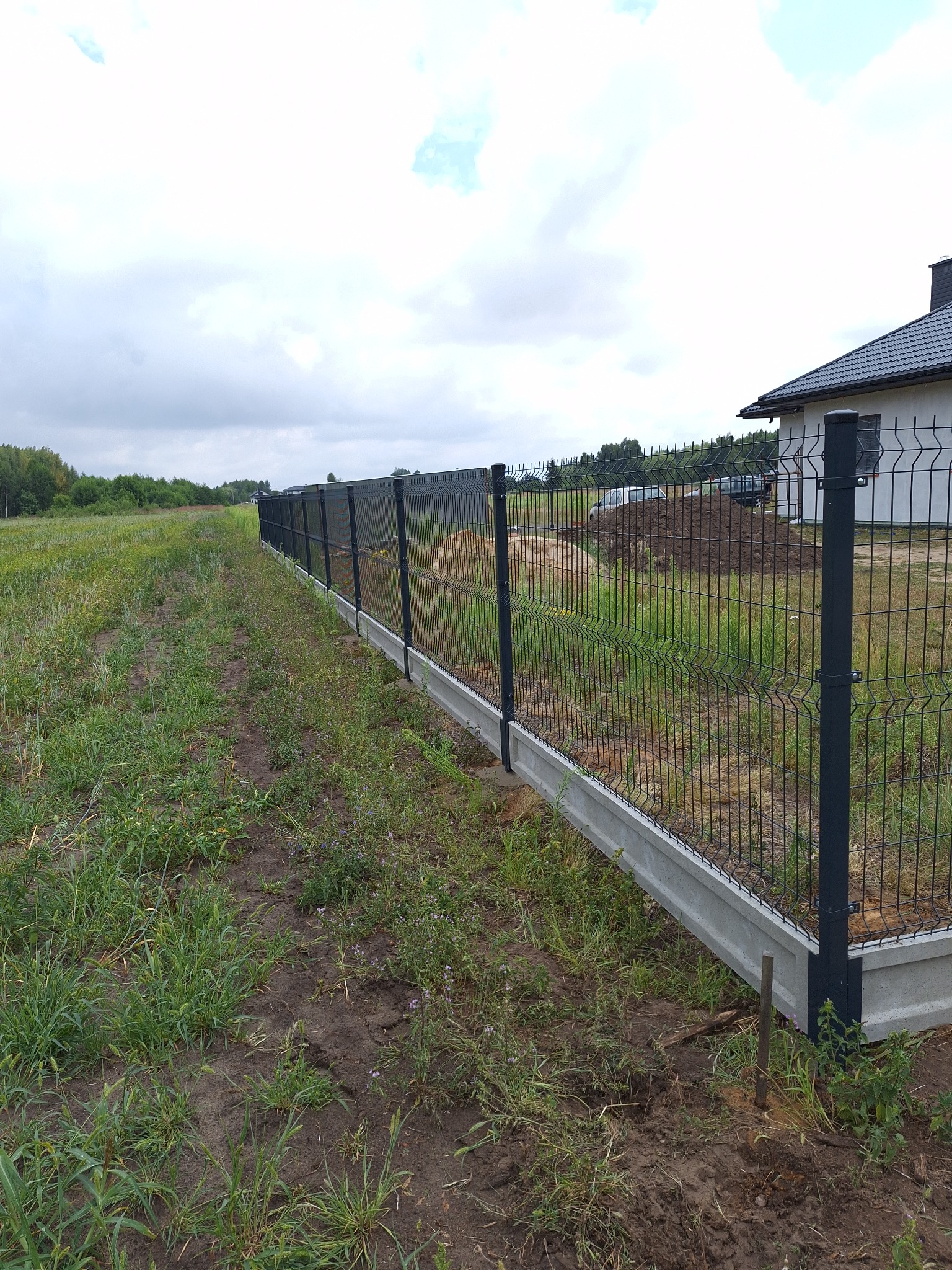 Długie, czarne ogrodzenie panelowe z podmurówką betonową, oddzielające posesję od łąki z chmurami w tle, widoczne świeże nasypy ziemi.