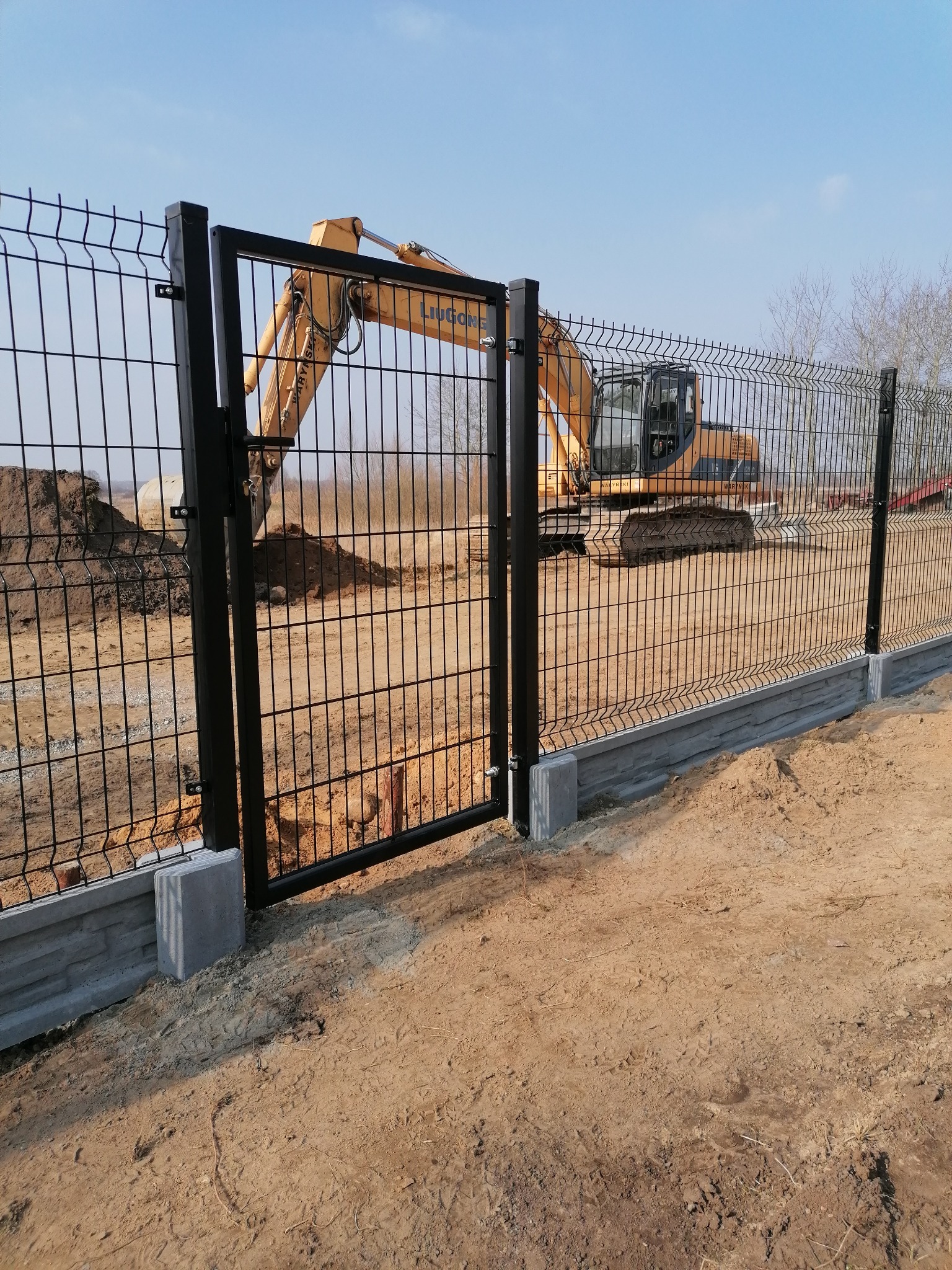 Nowoczesne ogrodzenie panelowe z czarną bramką i betonową podmurówką, w tle żółta koparka na placu budowy.