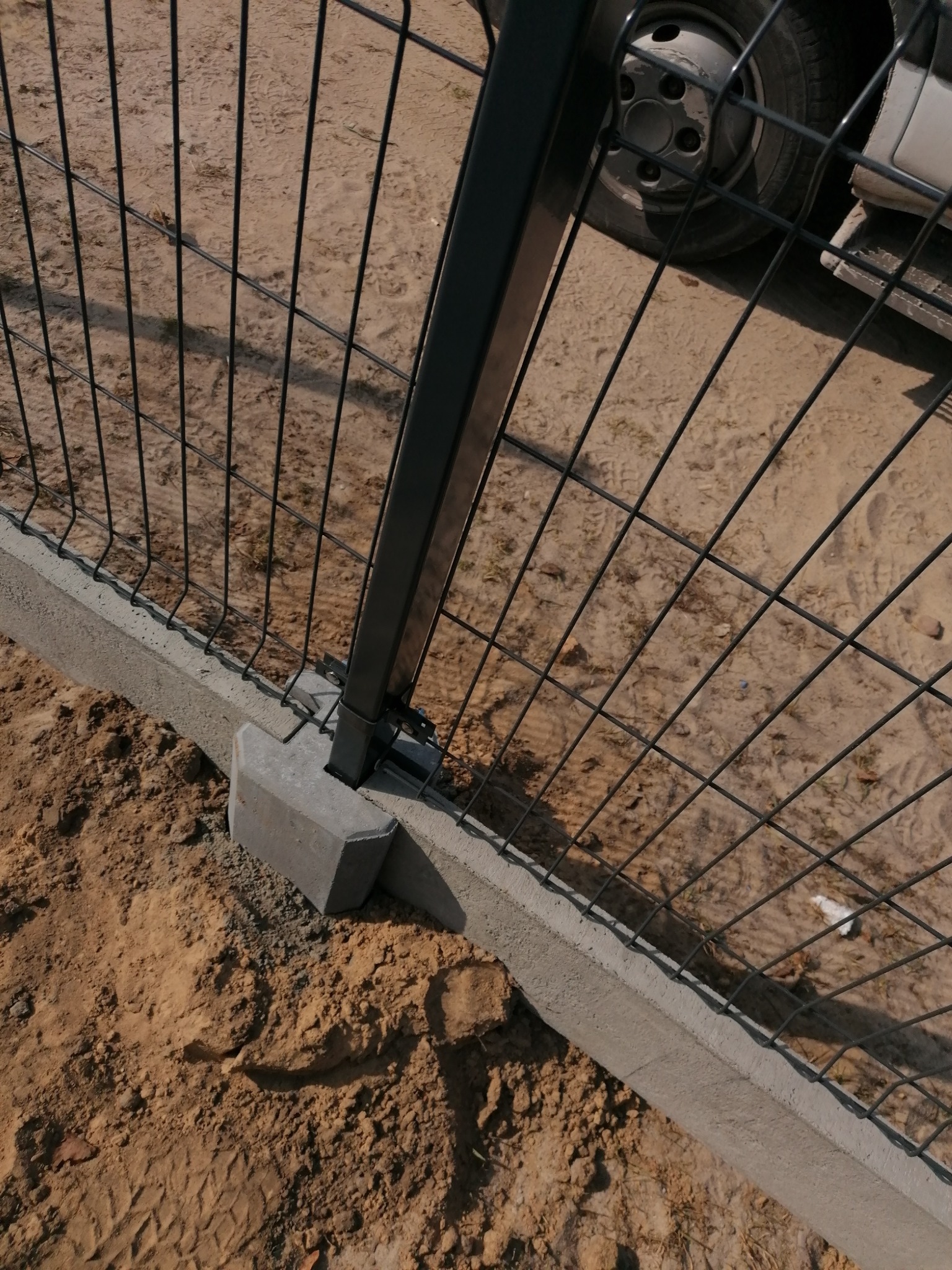 Fragment montażu ogrodzenia panelowego: czarny słupek z metalową siatką przymocowany do betonowej podmurówki na piaszczystym podłożu, widoczne ślady opon pojazdu w tle.