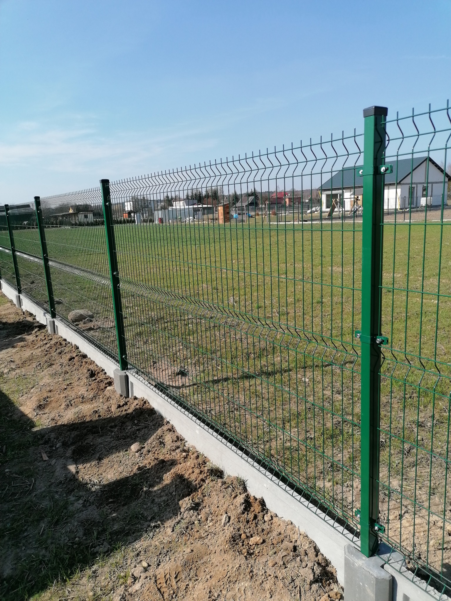 Fragment nowo zamontowanego, zielonego ogrodzenia panelowego z podmurówką betonową na tle trawnika i zabudowań jednorodzinnych w słoneczny dzień.