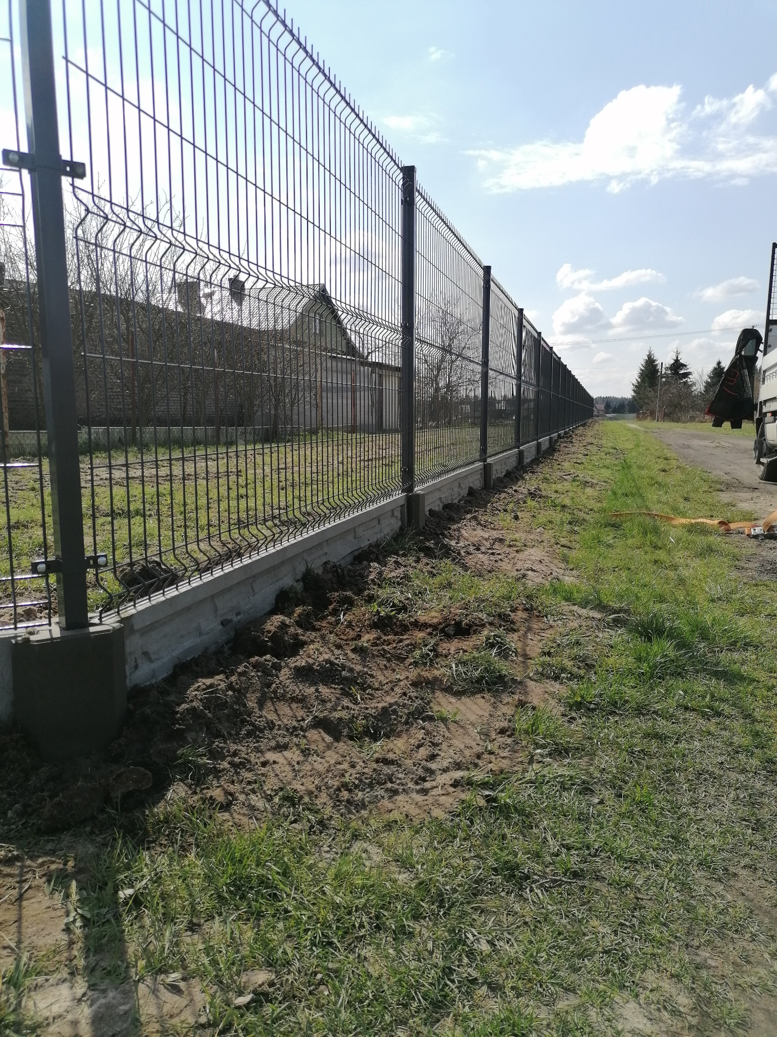 Długi, panelowy płot z podmurówką betonową, widziany z poziomu gruntu, z fragmentami ziemi i trawy po obu stronach, w tle dom i drzewa