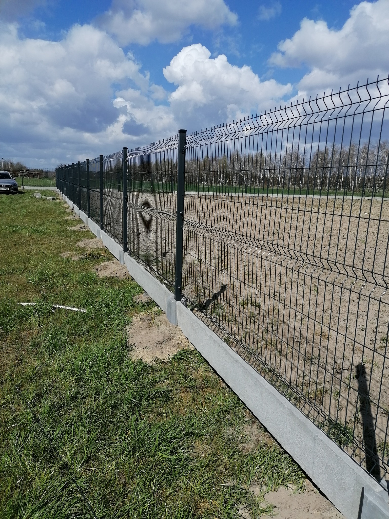 Długi fragment panelowego ogrodzenia z podmurówką betonową na trawiastym terenie, widok z boku w słoneczny dzień.