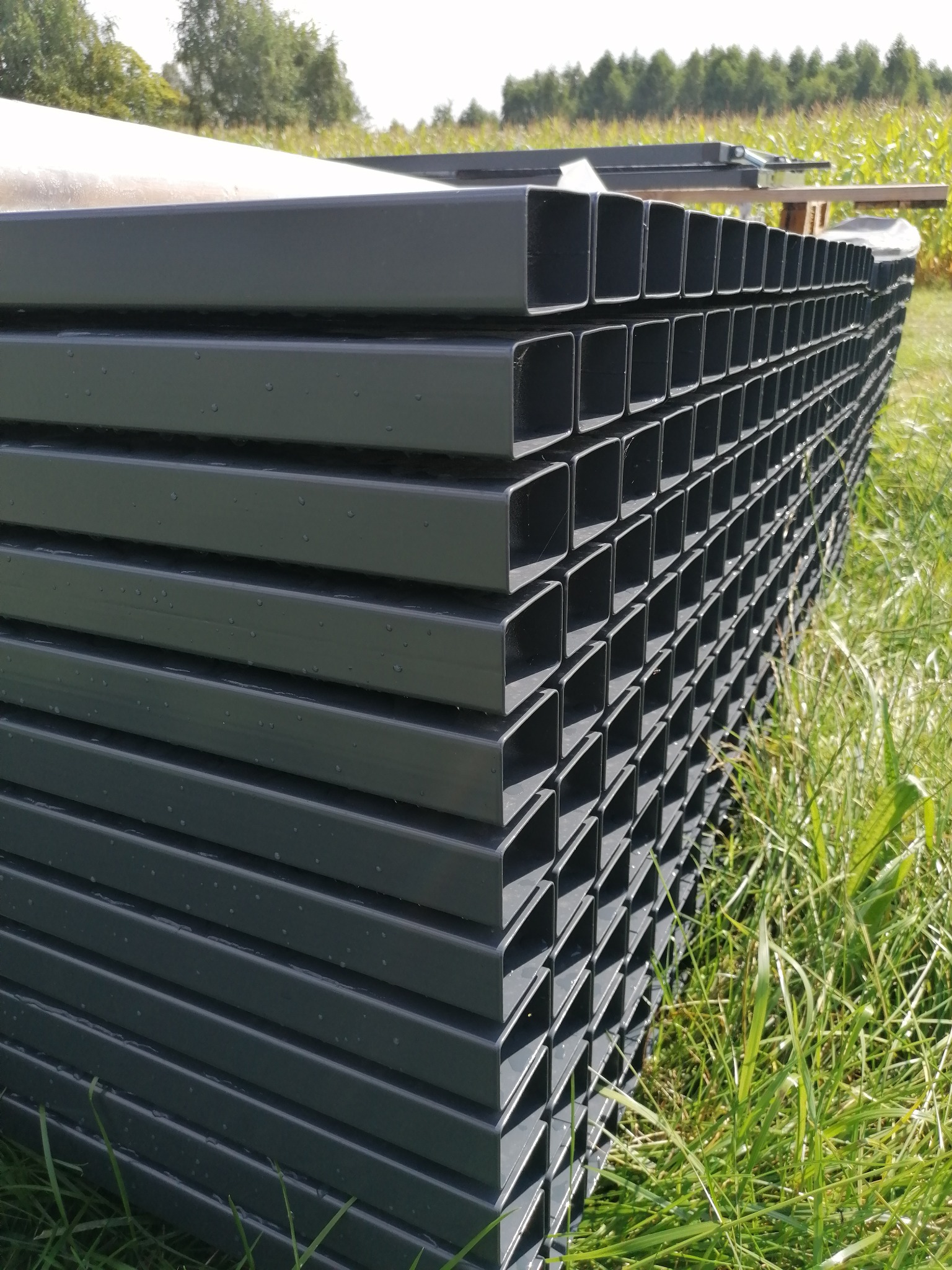 Stos szarych, kwadratowych profili metalowych ułożonych na trawie, przygotowanych do budowy ogrodzenia.