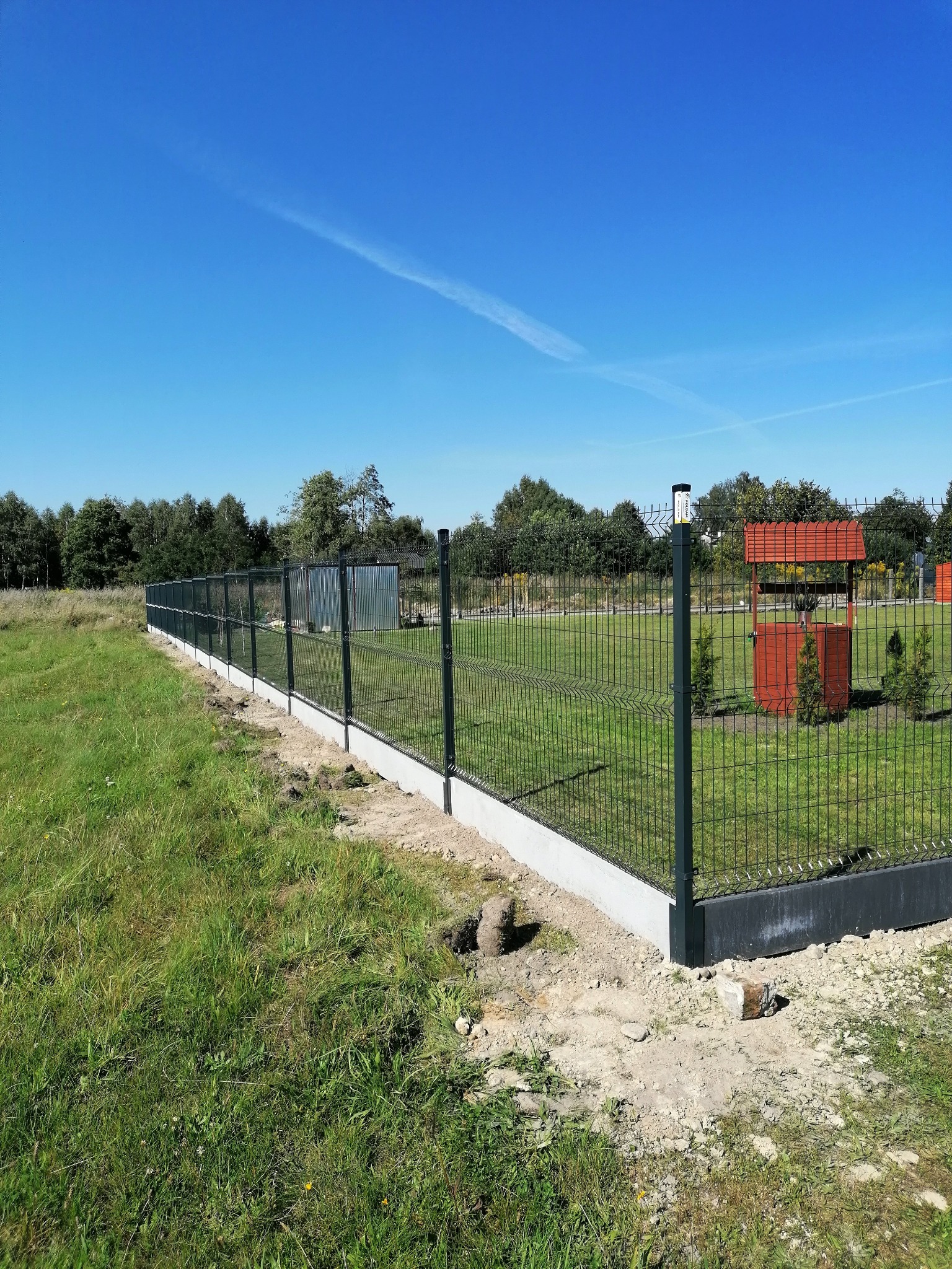 Długa, ciemnozielona siatka ogrodzeniowa na betonowej podmurówce, oddzielająca trawnik od terenu z drzewami i studnią z czerwonym dachem w tle, widok w słoneczny dzień.