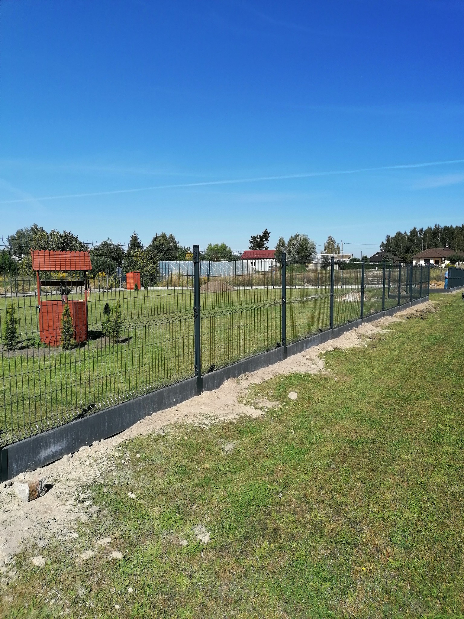 Nowoczesne ogrodzenie panelowe z podmurówką betonową, widoczne z bliska na tle zielonego trawnika i błękitnego nieba. W tle studnia zadaszona i inne elementy architektury ogrodowej.