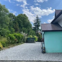 Ułożona kostka brukowa na podjeździe przed domem z turkusową elewacją, widoczna myjka ciśnieniowa i bujna roślinność w tle.