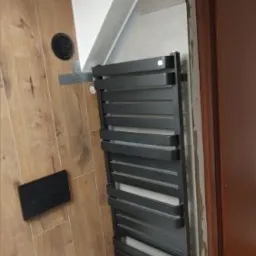 Nowoczesna łazienka w trakcie wykańczania: biała toaleta wisząca, czarny grzejnik drabinkowy na szarej ścianie, drewnopodobne płytki na ścianie z czarnym wentylatorem i przyciskiem spłuczki...