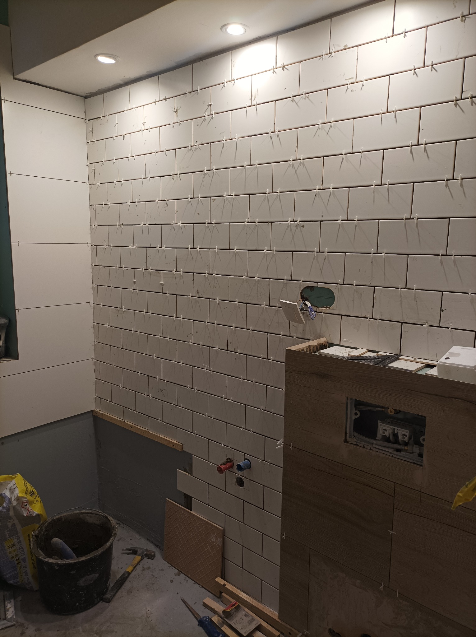 Białe, prostokątne płytki ułożone na ścianie łazienki, z widocznymi krzyżykami dystansowymi, częściowo zakryte drewnopodobną zabudową stelaża WC, z narzędziami i workiem zaprawy w tle.