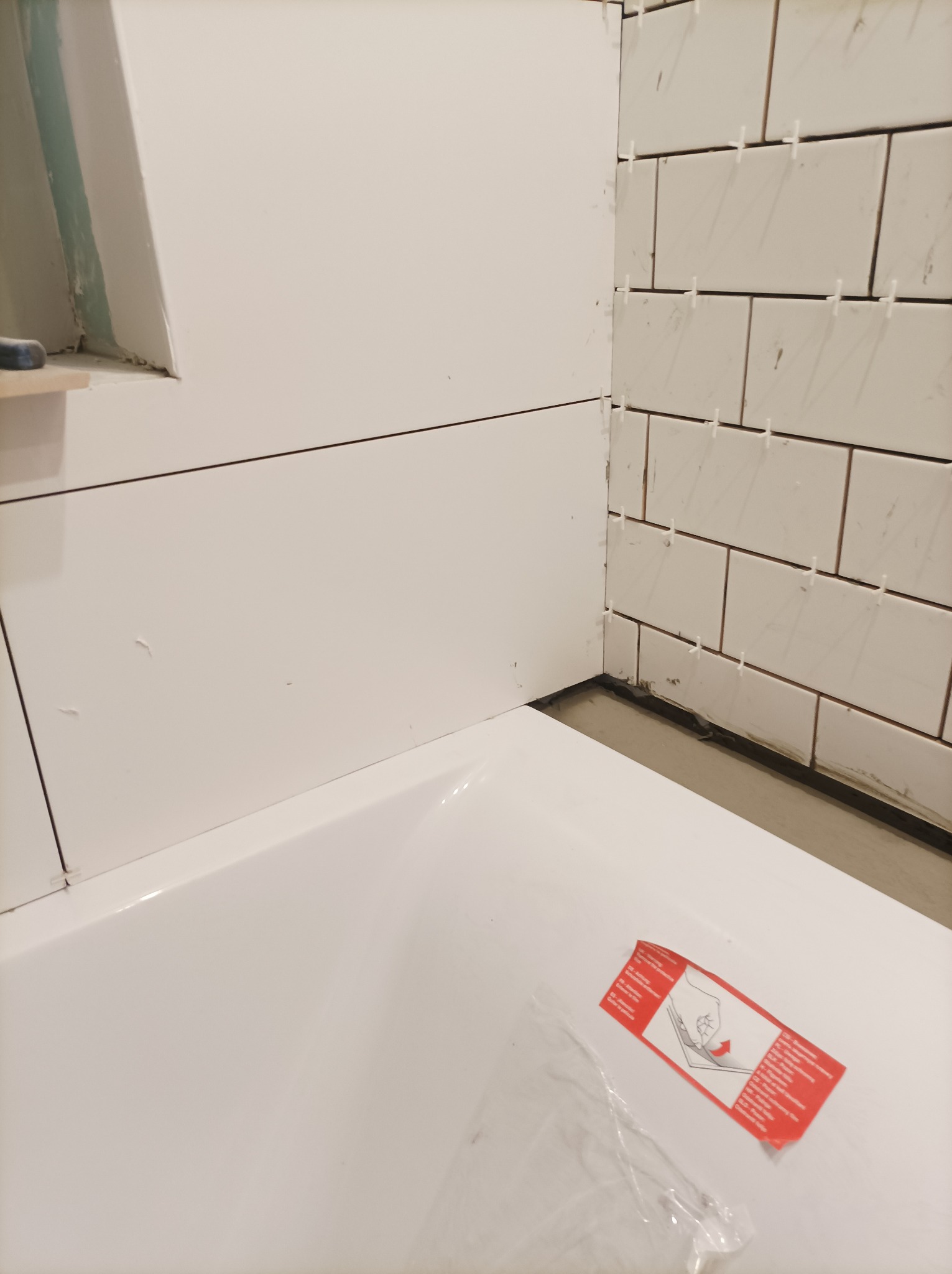 Biała wanna z częściowo wyłożonymi białymi płytkami, widoczne krzyżyki dystansowe, na wannie naklejka z instrukcją.