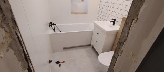 Wnętrze łazienki w trakcie remontu: biała wanna z czarną armaturą, umywalka z szafką w stylu retro, biała mozaika na ścianie i fragmenty ściany z widocznym klejem i zaprawą.