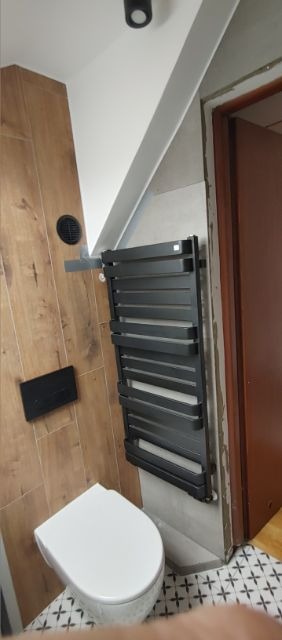 Nowoczesna łazienka w trakcie wykańczania: biała toaleta wisząca, czarny grzejnik drabinkowy na szarej ścianie, drewnopodobne płytki na ścianie z czarnym wentylatorem i przyciskiem spłuczki...