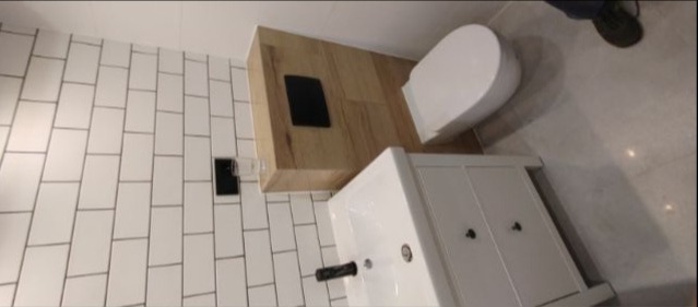 Nowoczesna łazienka z białą umywalką z szafką, wiszącą toaletą i białymi, prostokątnymi płytkami na ścianie, uzupełniona drewnopodobnymi panelami przy toalecie.