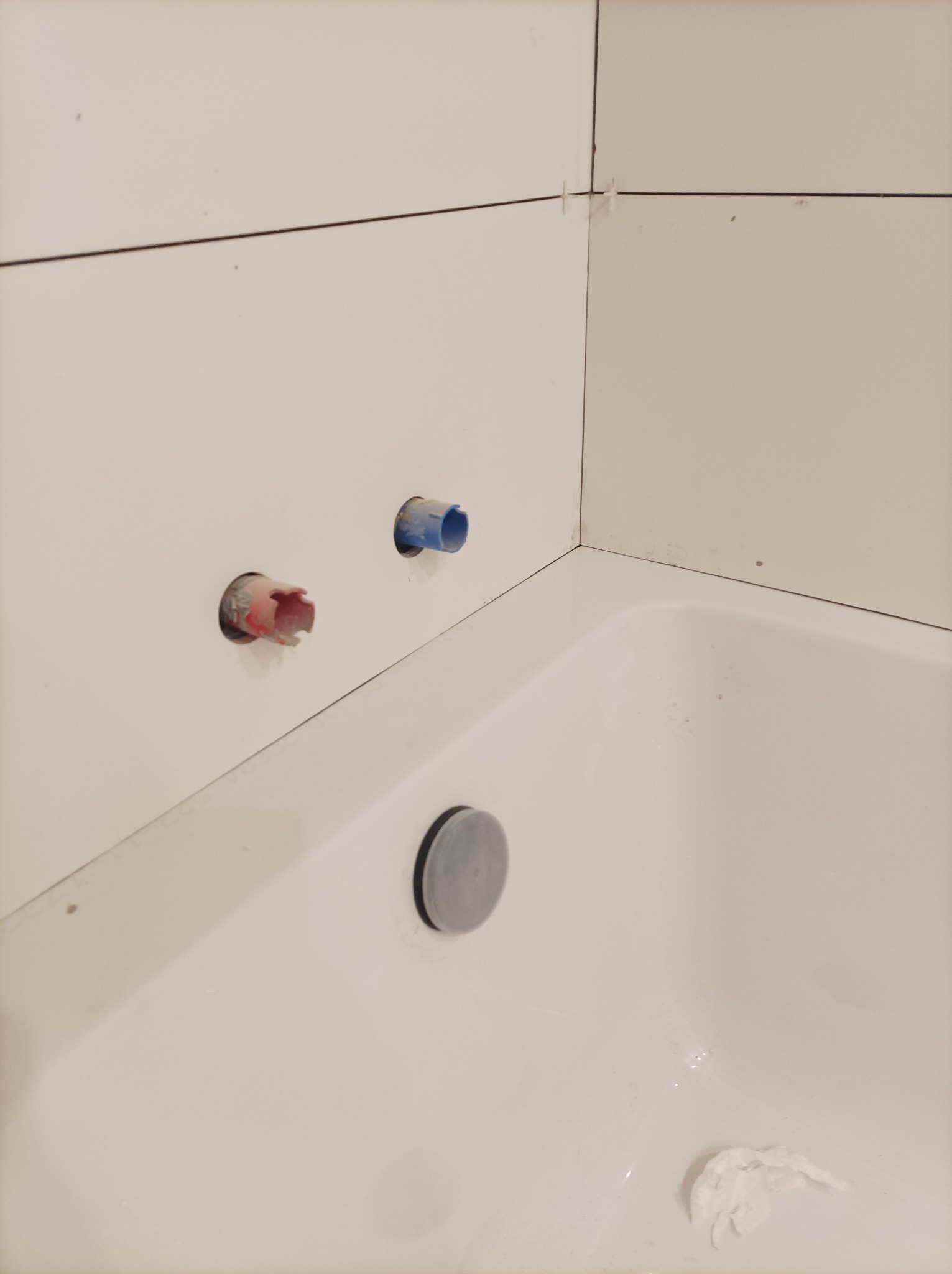 Biała wanna z widocznymi fragmentami świeżo ułożonych, białych płytek ściennych z krzyżykami dystansowymi, oraz wystającymi, zabezpieczonymi rurami instalacji wodnej.