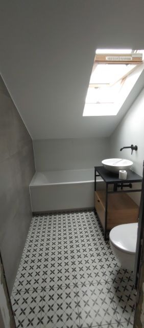 Wykończona łazienka na poddaszu ze skośnym sufitem i oknem dachowym, biała wanna, umywalka nablatowa na metalowym stelażu z drewnianą szafką, toaleta i wzorzysta podłoga.
