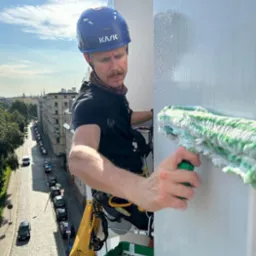 Pracownik w kasku KASK, zabezpieczony linami, myje okno na wysokości za pomocą zielonego mopa, widok na ulicę z perspektywy wysokościowej.