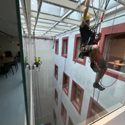 Dwóch pracowników na linach zabezpieczających czyści szklany dach wewnętrznego dziedzińca budynku, widoczne detale architektoniczne i odbicie w szkle.