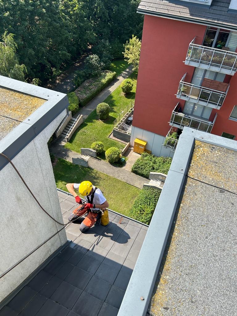 Pracownik w kasku i uprzęży na dachu budynku wielorodzinnego, zabezpieczony liną, używa narzędzia do obróbki dachu; w tle widok na zadbany ogród i elewację budynku.