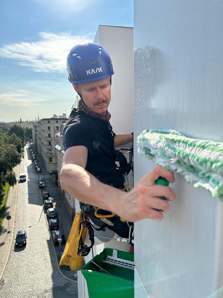 Pracownik w kasku KASK, zabezpieczony linami, myje okno na wysokości za pomocą zielonego mopa, widok na ulicę z perspektywy wysokościowej.