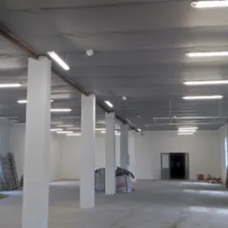 Wnętrze hali przemysłowej z białymi ścianami, kolumnami i rzędami świetlówek na szarym suficie podwieszanym, widoczne drabiny i materiały budowlane.