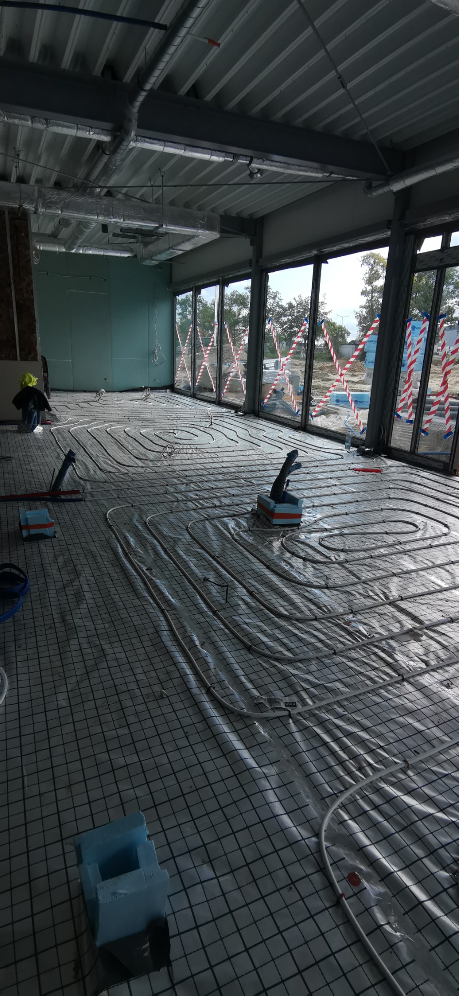 Montaż ogrzewania podłogowego w nowym budynku: białe rurki ułożone na folii z siatką, widoczne narzędzia i pracownik w tle.