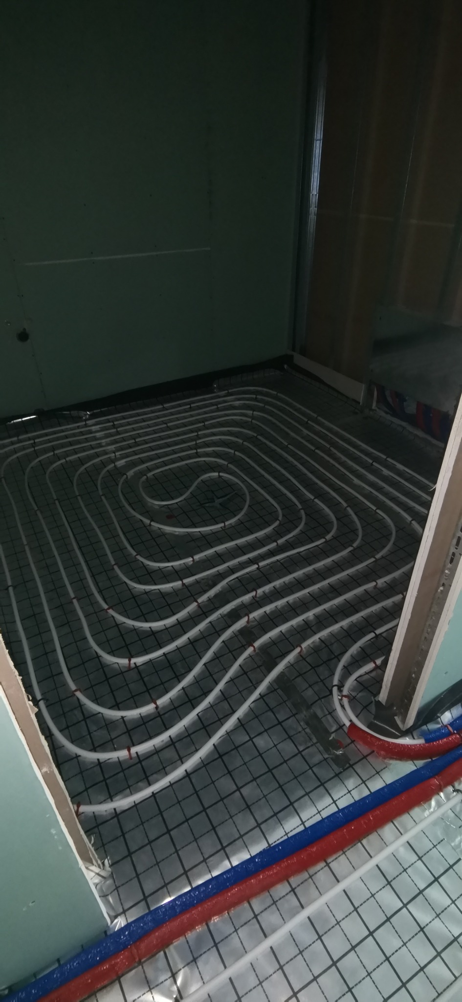 Układanie rur ogrzewania podłogowego na metalowej siatce, widoczne wejścia rur w kolorze czerwonym i niebieskim, ściany z płyt kartonowo-gipsowych w surowym stanie.