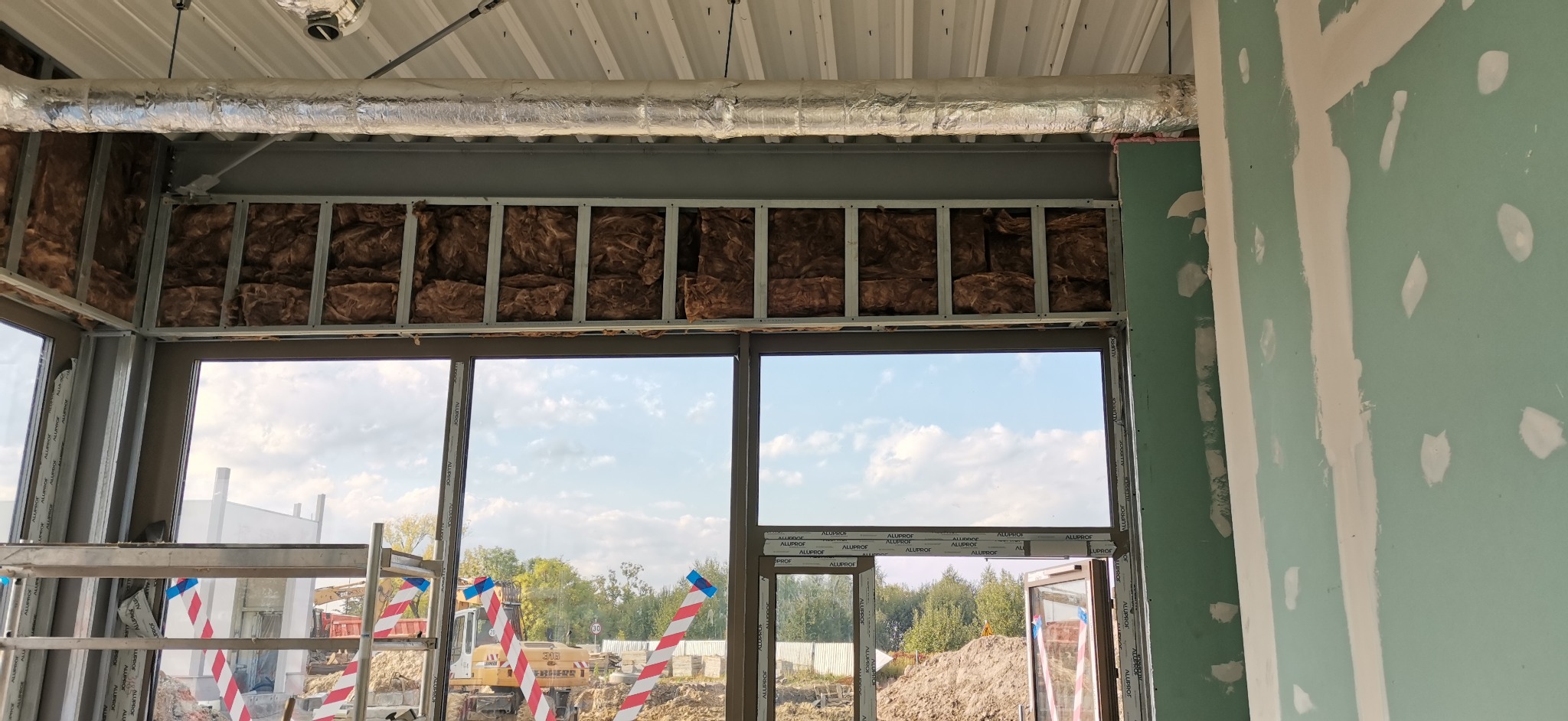 Montaż izolacji z wełny mineralnej w metalowym stelażu okiennym, widoczna płyta karton-gips na ścianie i rusztowanie w tle.
