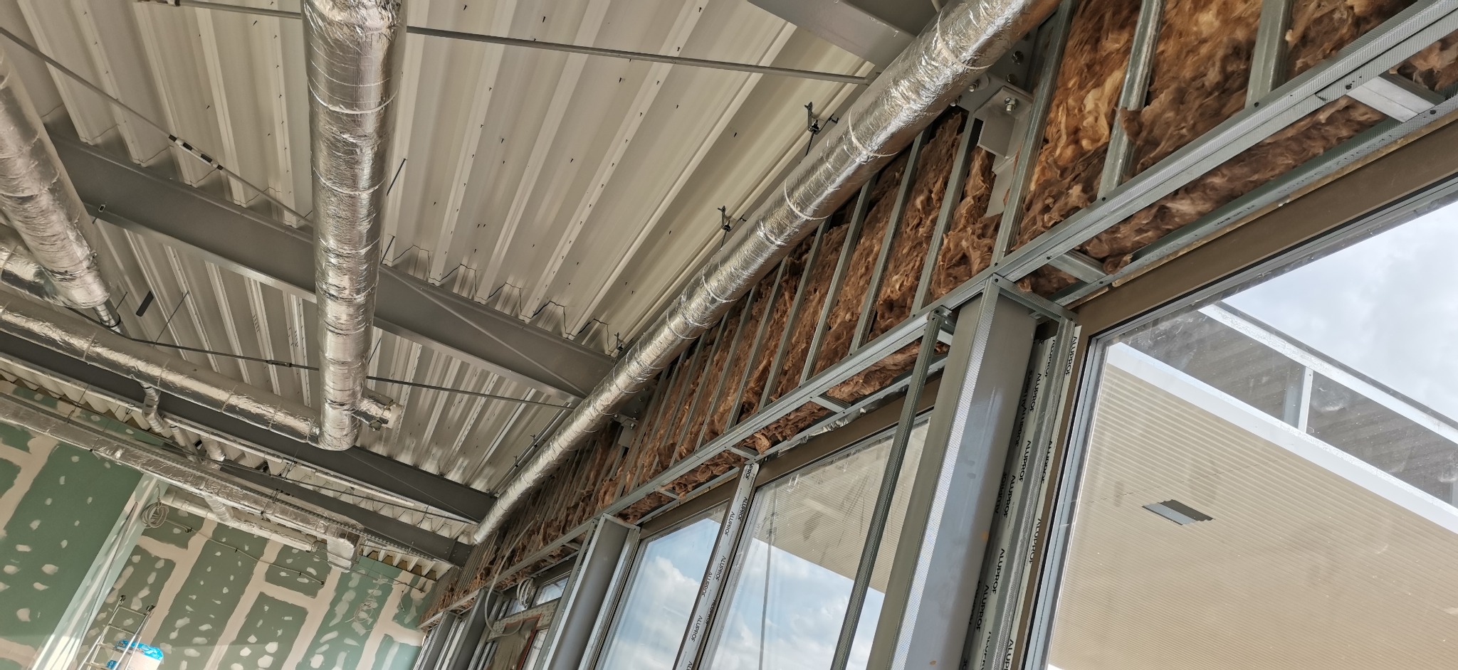 Montaż instalacji wentylacyjnej i izolacji w budynku w trakcie budowy, widoczne rury wentylacyjne z folii aluminiowej, metalowy stelaż pod płyty gipsowo-kartonowe oraz izolacja z wełny mineralnej.