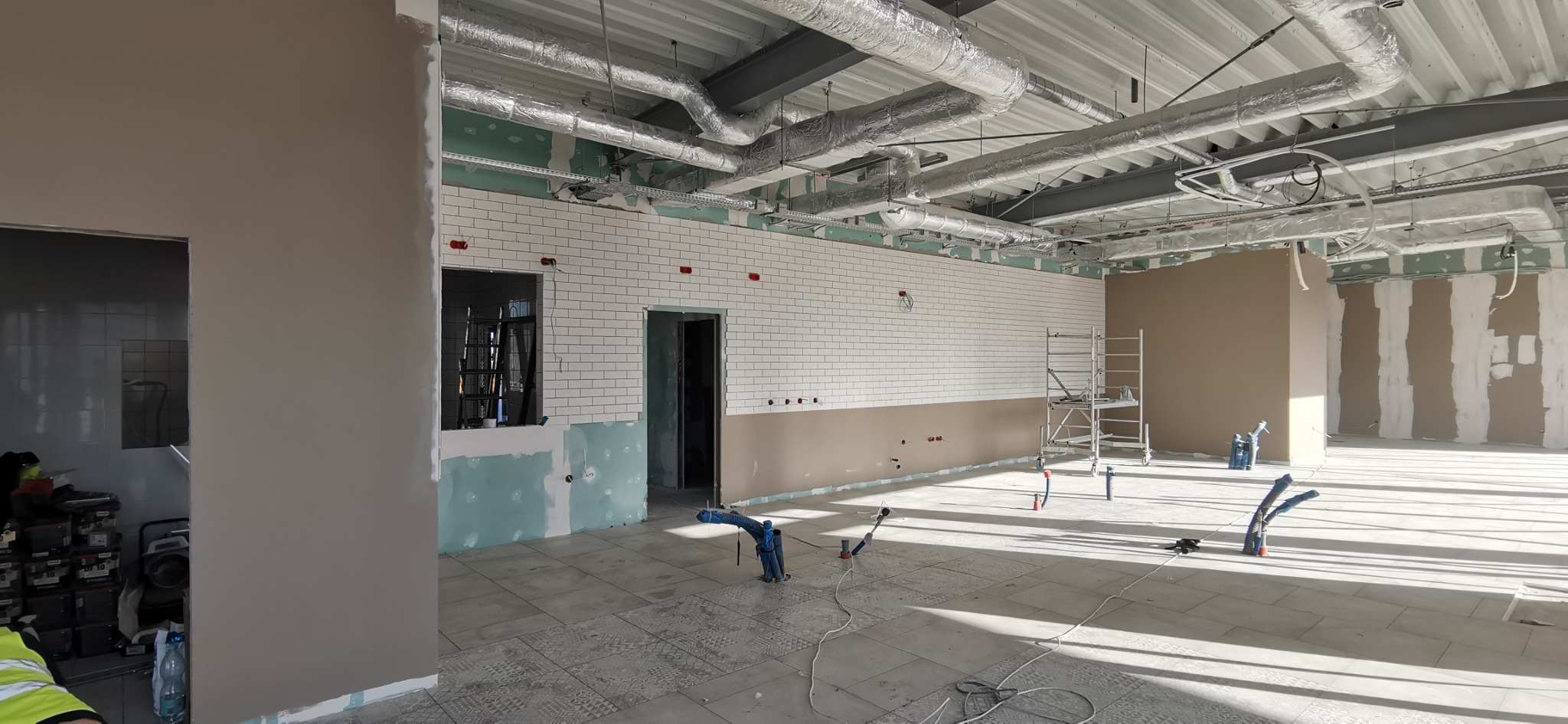 Wnętrze biura w trakcie prac wykończeniowych: ułożone białe płytki metra na ścianie, szpachlowane ściany gipsowe, widoczne rury wentylacyjne pod sufitem i rozłożone narzędzia na podłodze.