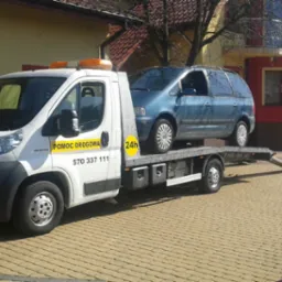 Biała laweta z napisem 'Pomoc Drogowa' i numerem telefonu przewozi niebieskiego minivana na tle brukowanej drogi i budynku z czerwoną fasadą.