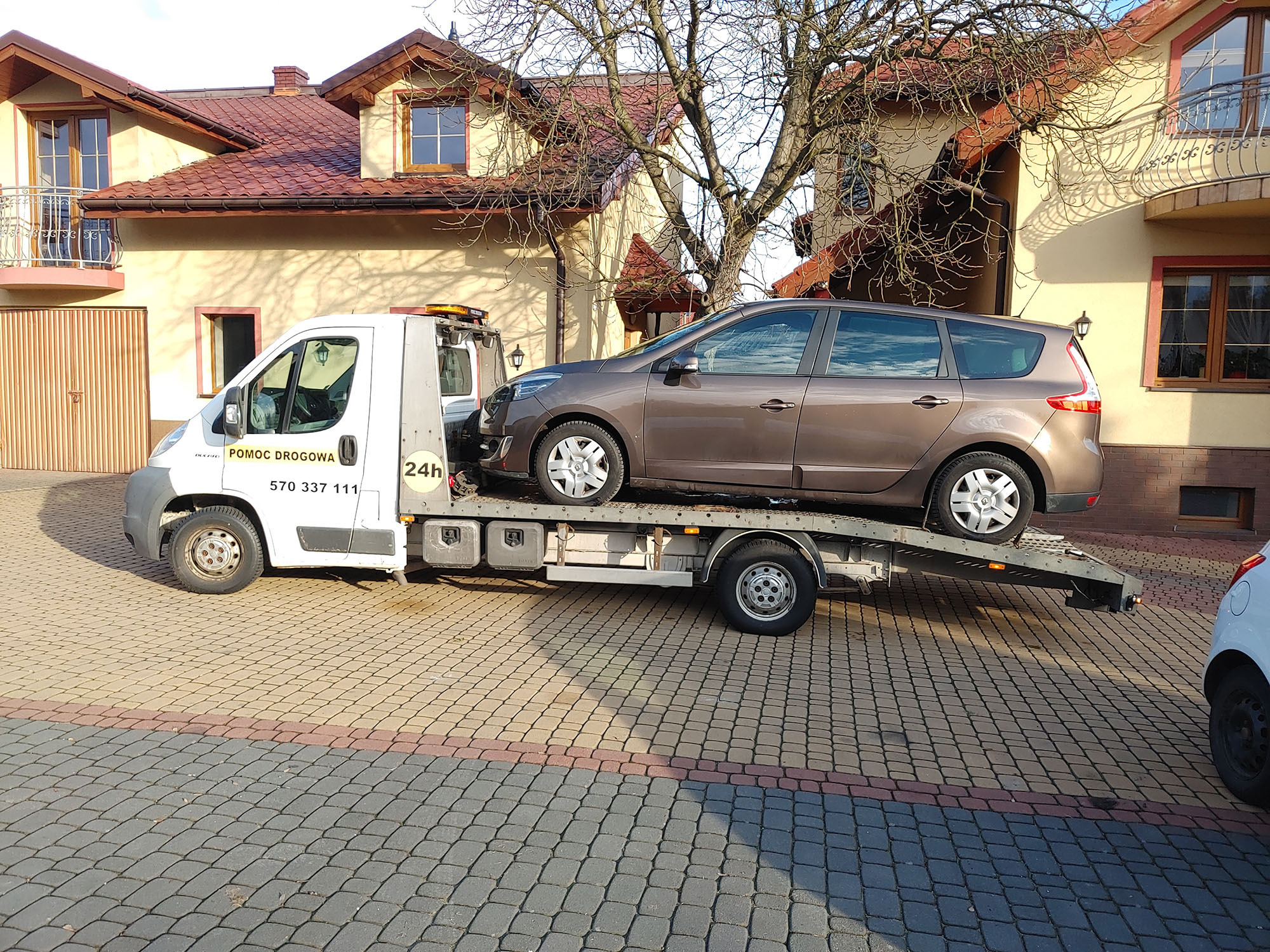 Brązowy samochód osobowy przewożony na lawecie pomocy drogowej przed budynkiem mieszkalnym.