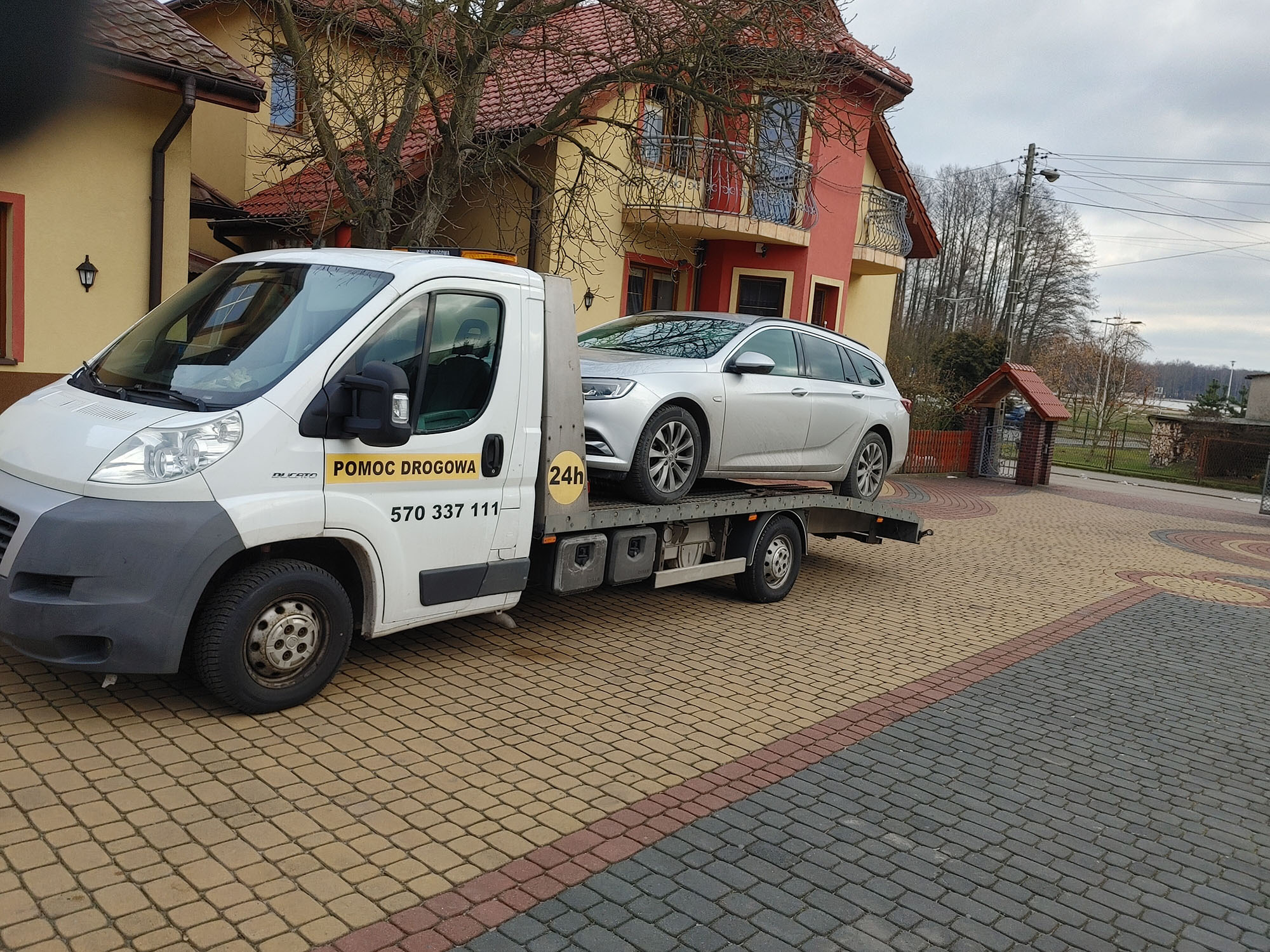 Biała laweta z napisem 'Pomoc Drogowa' i numerem telefonu przewozi srebrne kombi na tle kostki brukowej i dwupiętrowego domu z balkonem.