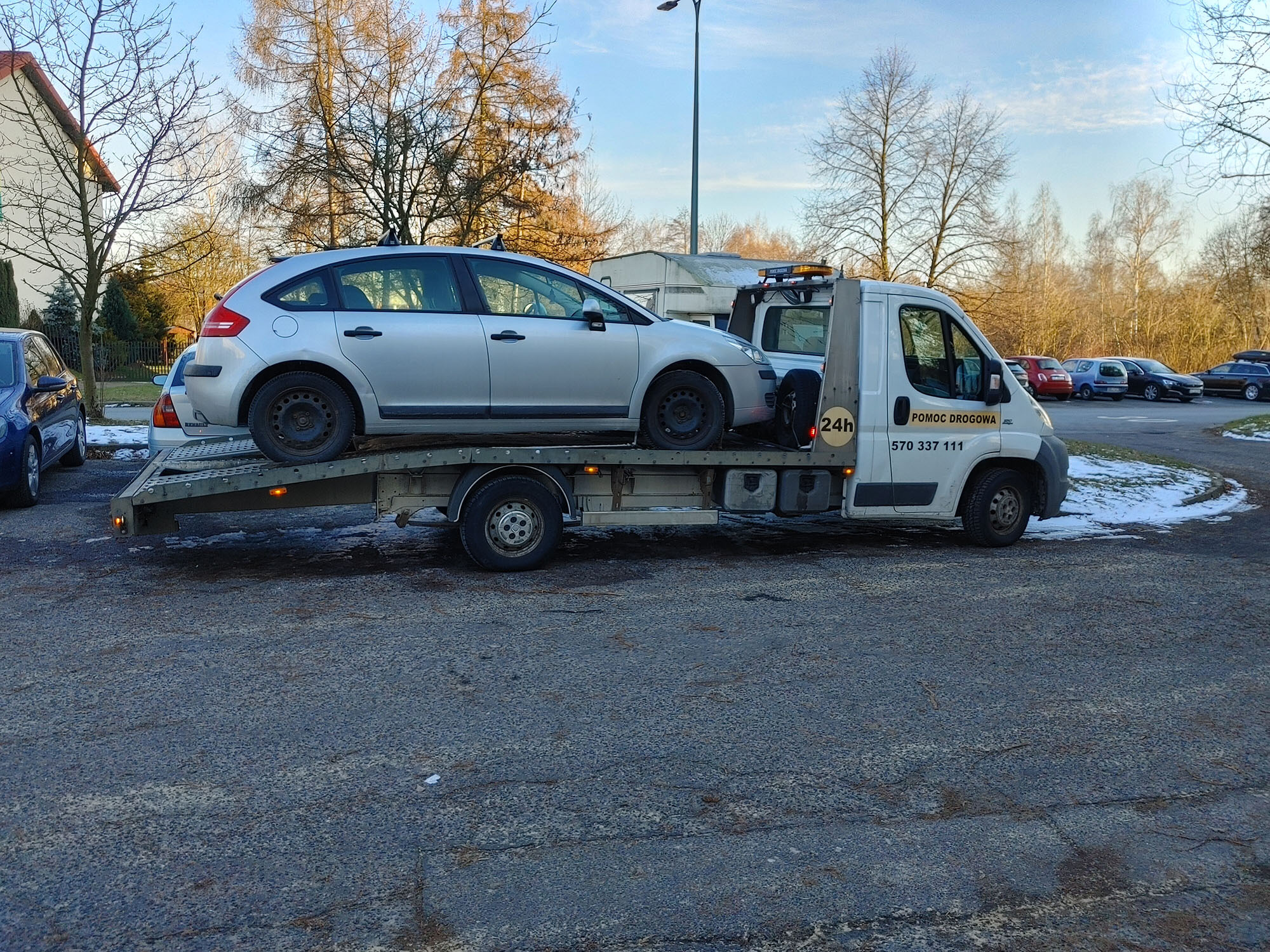 Srebrny samochód osobowy załadowany na platformę lawety pomocy drogowej z napisem '24h' na drzwiach, zaparkowanej na asfaltowym parkingu w słoneczny dzień.