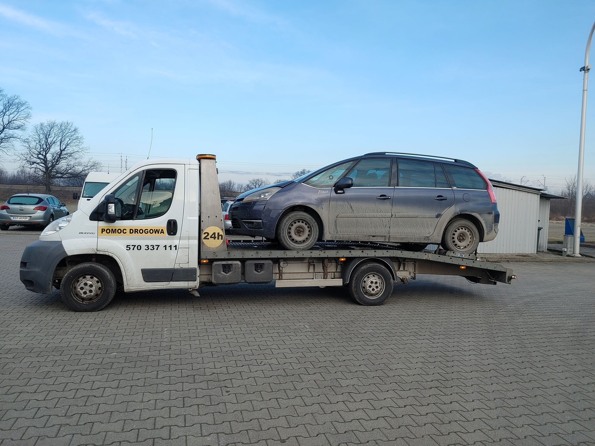 Biała laweta z napisem 'Pomoc Drogowa' i numerem telefonu przewozi brudny, granatowy samochód osobowy na tle szarej kostki brukowej i błękitnego nieba.