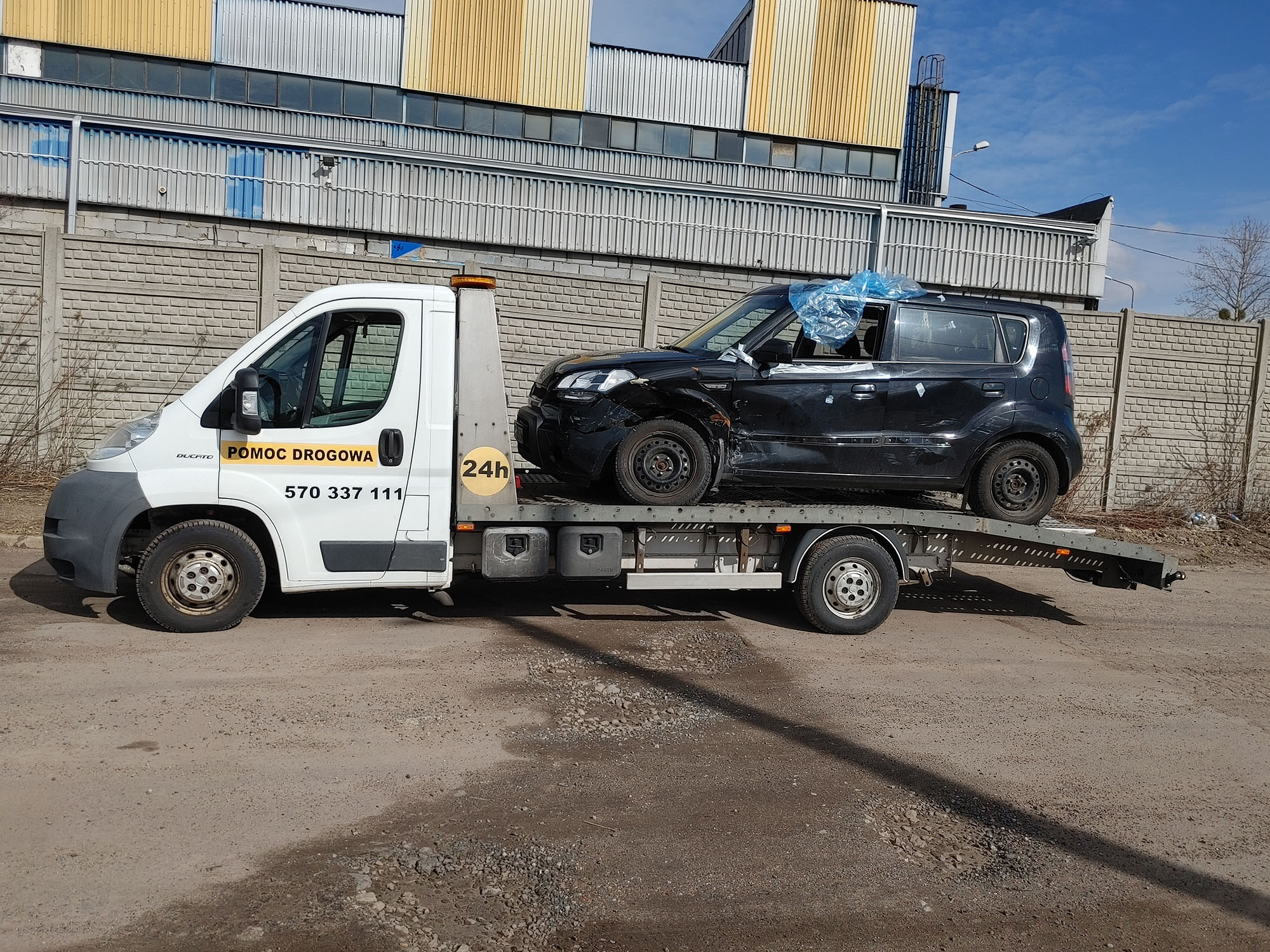 Czarny uszkodzony samochód osobowy zabezpieczony folią na lawecie białego pojazdu pomocy drogowej z numerem telefonu na tle szarego betonowego muru i hali przemysłowej w słoneczny dzień.