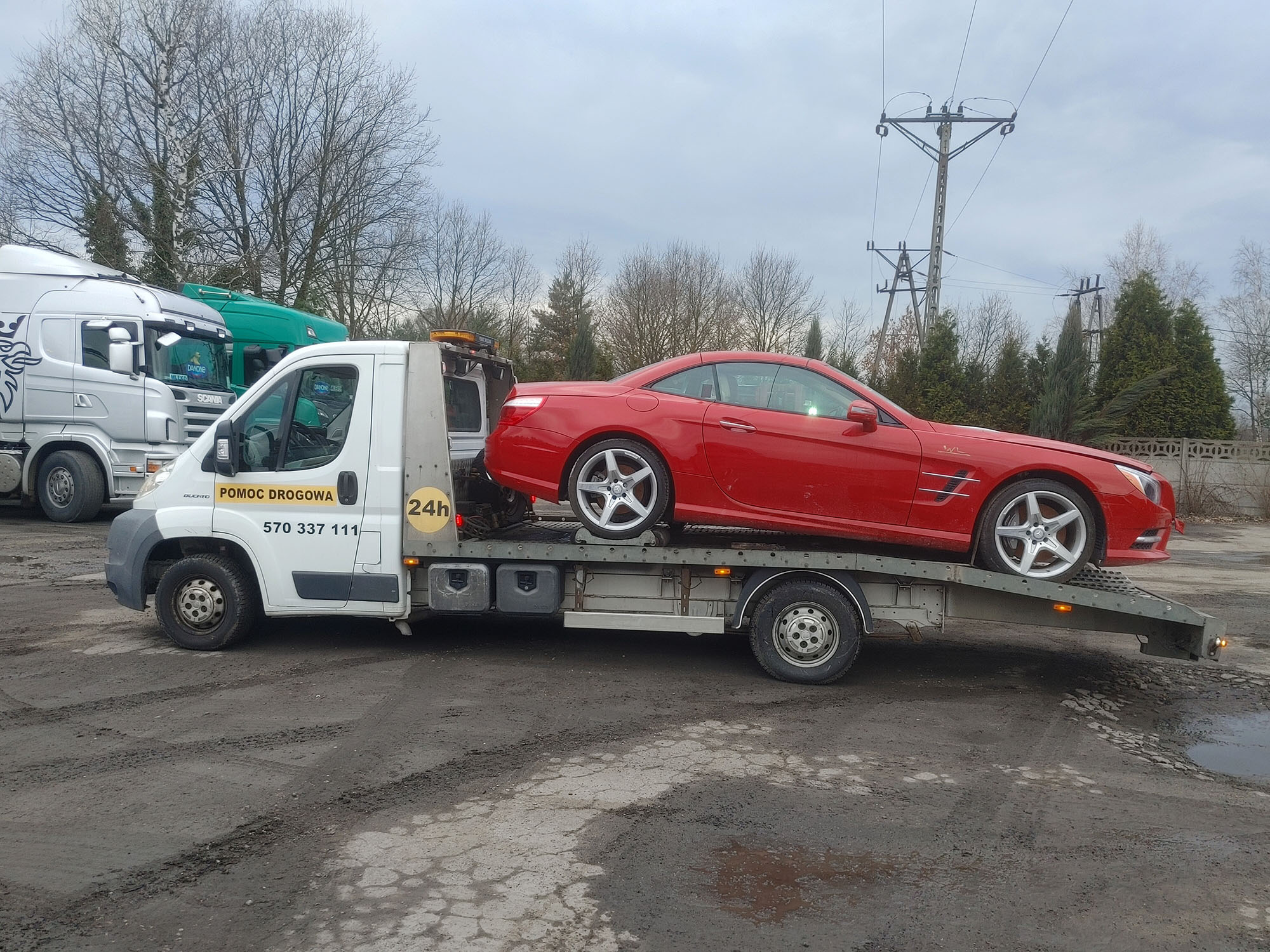 Czerwony kabriolet Mercedes na platformie białej lawety z napisem 'Pomoc Drogowa' i numerem telefonu, zaparkowanej na błotnistym placu, w tle inne ciężarówki i drzewa bez liści.