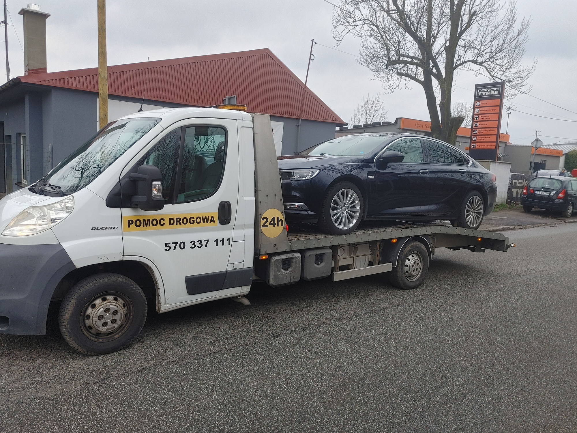 Czarny samochód osobowy na platformie białego pojazdu pomocy drogowej z żółtym napisem 'POMOC DROGOWA' i numerem telefonu, na tle budynku z czerwonym dachem i znaku reklamowego opon.
