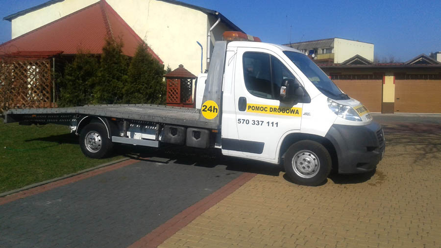 Biała laweta z napisem 'Pomoc Drogowa' na tle kostki brukowej i budynków.